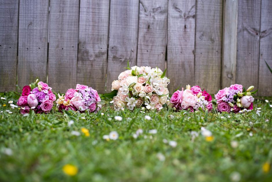 A line of five floral bouquets arranged on a grassy lawn in front of a weathered wooden fence. The bouquets feature various shades of pink, white, and cream flowers, including roses, hydrangeas, and f