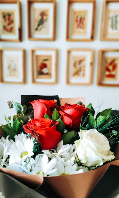 A close-up view of a floral arrangement featuring vivid red roses with velvety petals and fresh white daisies with yellow centers, complemented by lush green foliage. The bouquet is wrapped in light p