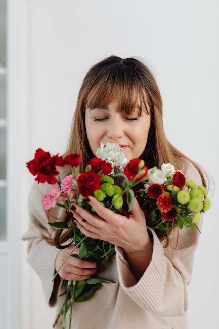 A woman with shoulder-length brown hair and bangs is holding a mixed bouquet of fresh flowers, including red carnations, pink and white roses, green chrysanthemums, and small red daisies, all arranged