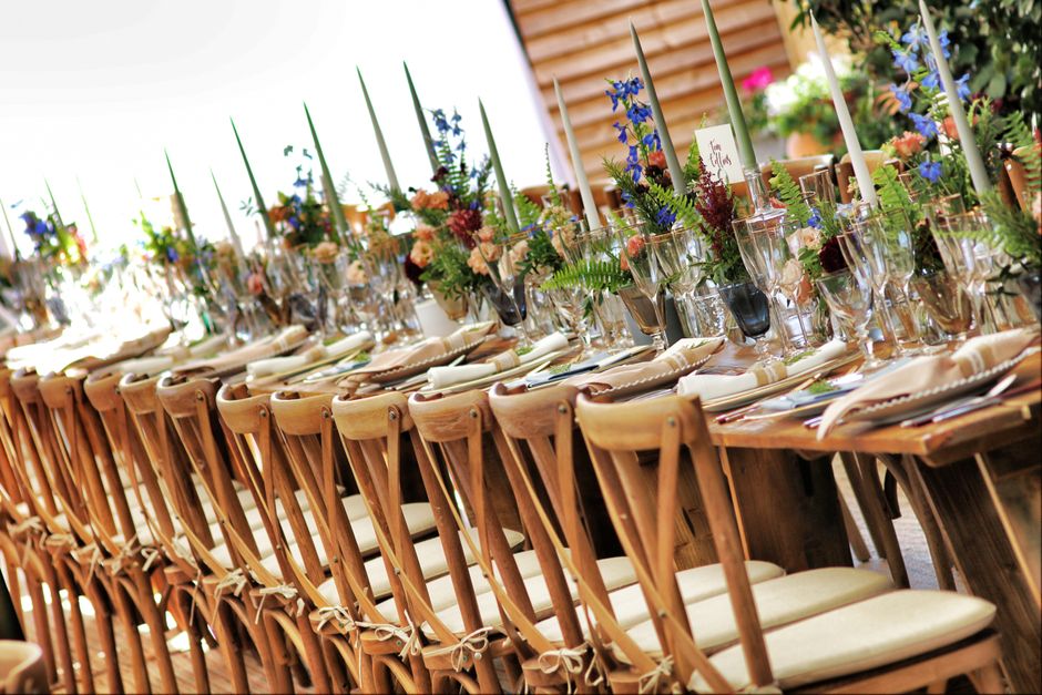 A long wooden banquet table set for a formal event with a floral arrangement featuring an assortment of fresh flowers in shades of pink, purple, blue, and cream, accented with green foliage. The table