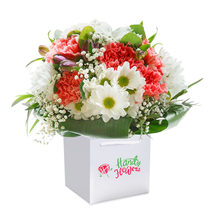 Expertly crafted by seasoned Petts Wood florists, this vibrant bouquet sits proudly in a flawless square white gift bag, presented against a pure white background to highlight its abundant beauty. The arrangement features clusters of soft coral-pink and luminous red carnations at their peak, their frilled petals forming a lively, textured tapestry. Among the bold carnations, crisp white daisy-like chrysanthemums with vivid yellow-green centers pop, their freshness bringing out memories of the blossom-filled corners of Jubilee Park. Ethereal sprigs of white baby's breath thread through the composition, softening transitions and lending an airy complexion to the bouquet. Near the lefthand side, discreet hints of purple-tinged buds-perhaps just-opening alstroemeria-peek through, drawing the eye with a soft promise of more blooms. Lush deep green leaves encircle the base, their sheen and generous form providing a natural, elegant collar that supports and elevates the seasonal palette. The sturdy white ribbon handles atop the bag connote convenience and thoughtful presentation, as if ready for delivery to a loved one's door along Queensway or Station Square. This bouquet radiates celebratory intent-ideal for birthdays, anniversaries, or simply to bring a measure of cheer into a family home in Petts Wood. Every visual element, from the interplay of light across the multi-textured petals to the crisp silhouette of the gift bag, expresses both freshness and the meticulous attention to detail that the local community has come to expect from its dedicated florists.
