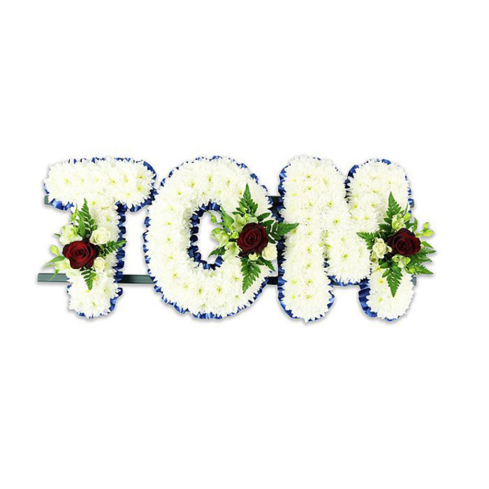 A poignant, handcrafted sympathy piece spells out TOM in large, sculptural letters, each letter densely clothed in countless small white chrysanthemum blooms that give the surface a soft, tactile fullness like velvet under the fingertips. The precise edges of every letter are neatly finished with a vivid royal blue ribbon, its satiny line providing a crisp graphic contrast that frames the white field and makes the name legible at a glance. Set into the centre or lower area of each character is a formal spray: a solitary deep red rose anchors the cluster, its rich hue and velvety texture the emotional heart of the design, while tiny creamy-white spray roses and slender fern fronds are artfully arranged around it to add lightness, leafed texture and a muted green punctuation. The roses bring a subtle, rounded perfume while the chrysanthemums remain quietly neutral, together producing an atmosphere of respectful calm. The letters are supported by a slim, dark green or black metal framework and a discreet horizontal bar visible behind the base, a practical touch that keeps the tribute upright whether placed on easels at a chapel or laid beside a casket at a service near Petts Wood Common. Rendered against a clean white backdrop and photographed in soft, diffused daylight, this arrangement communicates dignity and personalised care-a classic memorial aesthetic thoughtfully created to honour a life among friends and family in Petts Wood and the surrounding borough.