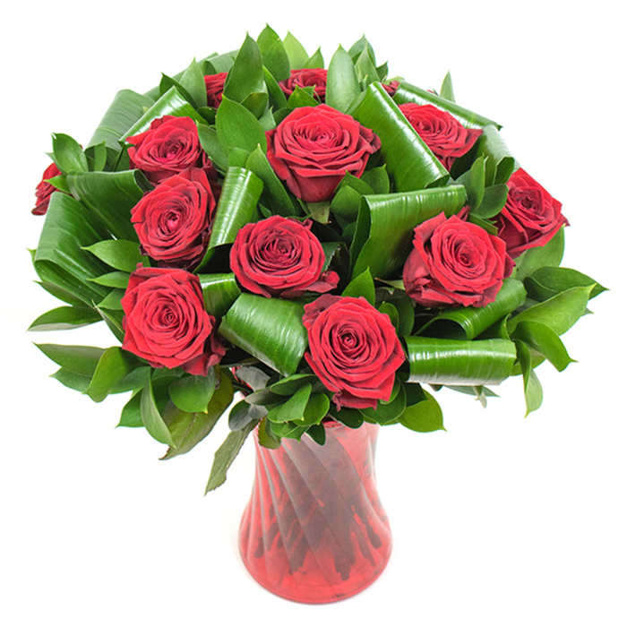 A carefully composed bouquet of roughly a dozen deep crimson roses takes centre stage, every bloom fully open with tightly spiralled centres that ease into soft, velvety outer petals. The rich red of the roses is deep and saturated, offering a classic declaration of love that reads timeless in the even studio lighting. Interspersed among the blooms is lavish dark green foliage: narrower, glossy leaves form a dense, supportive backdrop while larger, glossy green leaves have been artfully rolled and shaped into cylindrical curls that curve around individual rose heads, introducing a contemporary, sculptural rhythm to the arrangement. The florist's technique is clear in the balanced volume and the crisp way each element breathes within the composition. The bouquet is presented in a tall, slender ruby-red glass vase with a faintly ribbed, twisted texture; its translucent surface catches and reflects light, echoing the vertical stems and lending the piece a refined, jewel-like presence. Placed against a stark white background, the colours pop with gallery-like clarity, making the arrangement an elegant option for anniversaries, Valentine's Day, or a romantic surprise. As prepared locally in Petts Wood by Flower Delivery Petts Wood, this design exemplifies neighbourhood craftsmanship - ideal for same-day delivery around Petts Wood village, the station, and nearby Bickley or Orpington when a statement of enduring devotion is needed.
