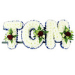 Shown here is a personalised funeral flower arrangement spelling out the name TOM in large, upright capital letters, ideal for a memorial or graveside tribute in the Petts Wood area. Each letter is meticulously packed with white chrysanthemum blooms, arranged so densely that they create a smooth, plush surface, like a soft floral cushion. The petals are crisp and fresh, giving the impression of flowers recently arranged by a skilled local florist. Encircling every letter is a royal blue satin ribbon, gently pleated and pinned into place, its glossy finish catching the light and defining the shape of each character against the clean white backdrop of the photograph. At three main points-on the T, O, and M-carefully composed floral sprays add richness and colour. These sprays feature deep red roses with velvety, layered petals, interspersed with smaller cream or white spray roses and slim white blooms reminiscent of freesia, all surrounded by a collar of fresh green foliage and delicate fern fronds. The interplay of vivid red, serene white, cool blue, and natural green creates a harmonious palette that speaks of love, respect, and peace. The letters are mounted along a narrow dark rail, ready to be placed on a stand, hearse, or casket for a service at a local church or at one of the crematoriums serving Petts Wood. Overall, the tribute feels both traditional and deeply personal, a floral memorial designed to honour the life and memory of someone dearly loved named Tom.