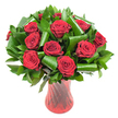A lush, rounded bouquet of a dozen-plus deep crimson roses fills the frame, each bloom at the height of perfection with velvety petals spiralling from a tight centre to gently unfurling outer layers. The colour is almost wine-dark in places, a passionate red that reads classic yet modern under soft, even studio light. Interwoven between the rose heads is a generous, sculptural mass of glossy dark green foliage: flat, leathery leaves that form a dense verdant backdrop and broader, emerald-green leaves skillfully folded and rolled into cylindrical loops that arch and cradle the flowers like living architecture. The contrast between the smooth, reflective surfaces of the leaves and the plush, matte texture of the rose petals gives the arrangement depth and a tactile richness you can almost feel. Everything sits in a tall, slender ruby-red glass vase whose translucent surface carries a subtle ribbed or twisted motif; vertical lines catch the light and echo the stems' upward movement, while the vase's narrow waist and flared mouth provide a graceful frame for the blooms. Set against a pure white background, the red and green tones pop with gallery clarity, turning the bouquet into a striking focal point for an anniversary table or a romantic centrepiece. As a Petts Wood florist might describe it, this arrangement balances traditional romance with contemporary leafwork, evoking the quiet charm of the village green and the warm neighbourhood moments that make local celebrations memorable.