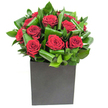 In this elegantly modern arrangement, a cluster of rich, velvety crimson roses takes centre stage, each bloom opened to reveal soft, spiral layers of petals that read as both intimate and dramatic. The roses are nestled in a choreography of broad, glossy green leaves that have been carefully curled and looped to create a sense of movement and rhythm; their waxy sheen catches the light and frames each red bloom like a living silhouette. The composition sits low and confident in a matte black cube vase, its geometric form grounding the design with a contemporary edge that feels equally suited to a city flat or a polished office reception. Lighting is clear and steady, lifting tiny highlights off petal edges and the satiny surfaces of the foliage so the crimson appears almost tactile. Close your eyes and you can imagine a delicate perfume of classic rose softened by the cool, green notes of freshly cut leaves, a scent that reads as clean and uplifting rather than cloying. The overall mood balances passion with restraint-a bouquet made to mark anniversaries, heartfelt apologies, or to bring warm, romantic drama to a modern dining table. As a local florist I picture this arrangement brightening a window seat near Petts Wood station or taking pride of place on a mantel in a home just off the village green; it is crafted with attention to proportion and longevity, designed to arrive fresh and to make a memorable impression without overstating itself.